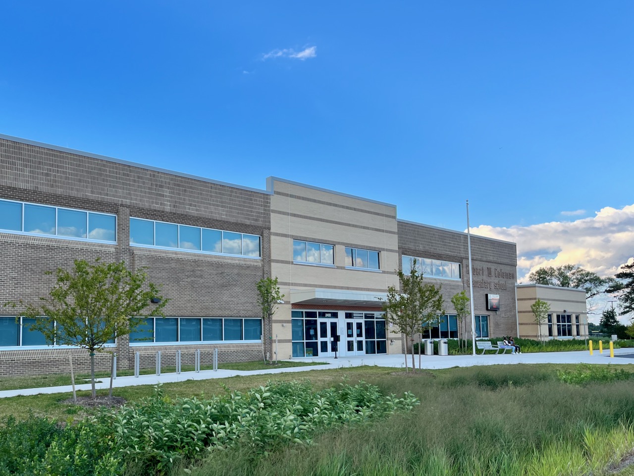 View of a two-story brick school building with a large modern entrance covered by a projecting awning. A rain garden appears in the foreground.