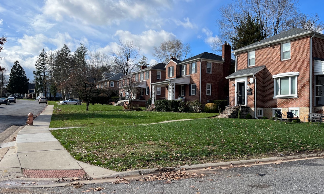 A view of a of brick detached houses with wide front lawns, on the right, and a concrete sidewalk next to the street, on the left.